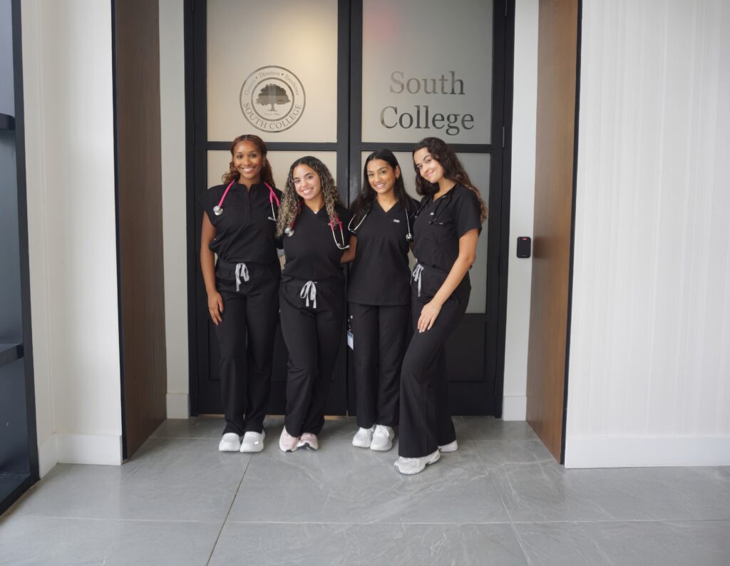 Four women in black scrubs and white shoes stand smiling together in front of glass doors labeled "South College." Two of them have stethoscopes around their necks. They are PA students.