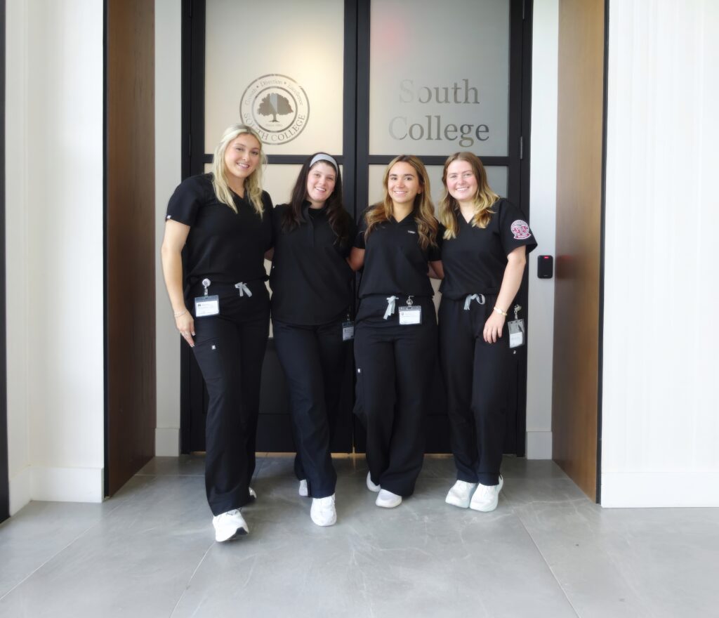 Four women in black scrubs and white shoes stand smiling together in front of a glass door labeled "South College." They each wear name badges.