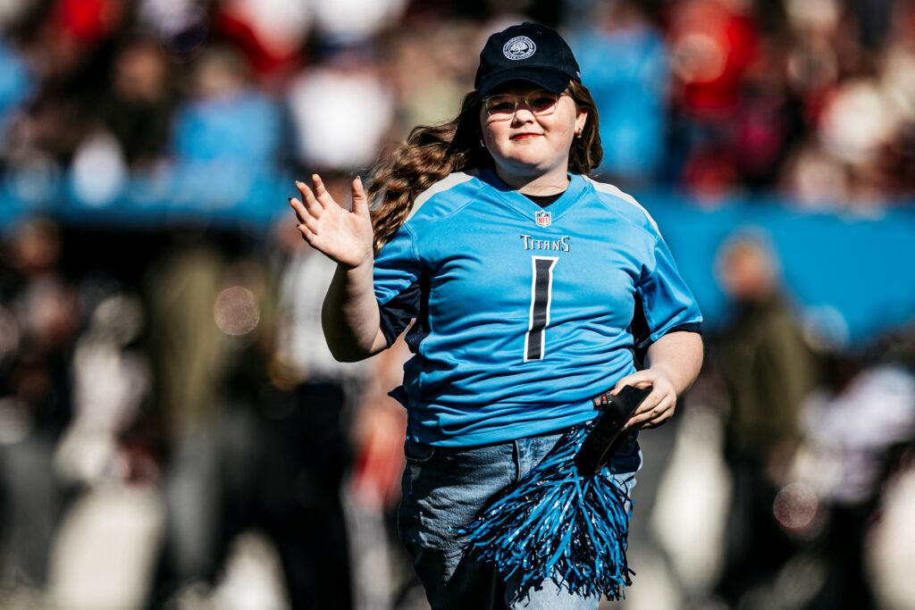 A girl in a blue Tennessee Titans jersey and cap waves and smiles while holding pom-poms on a football field. The background shows a blurred crowd and other people.