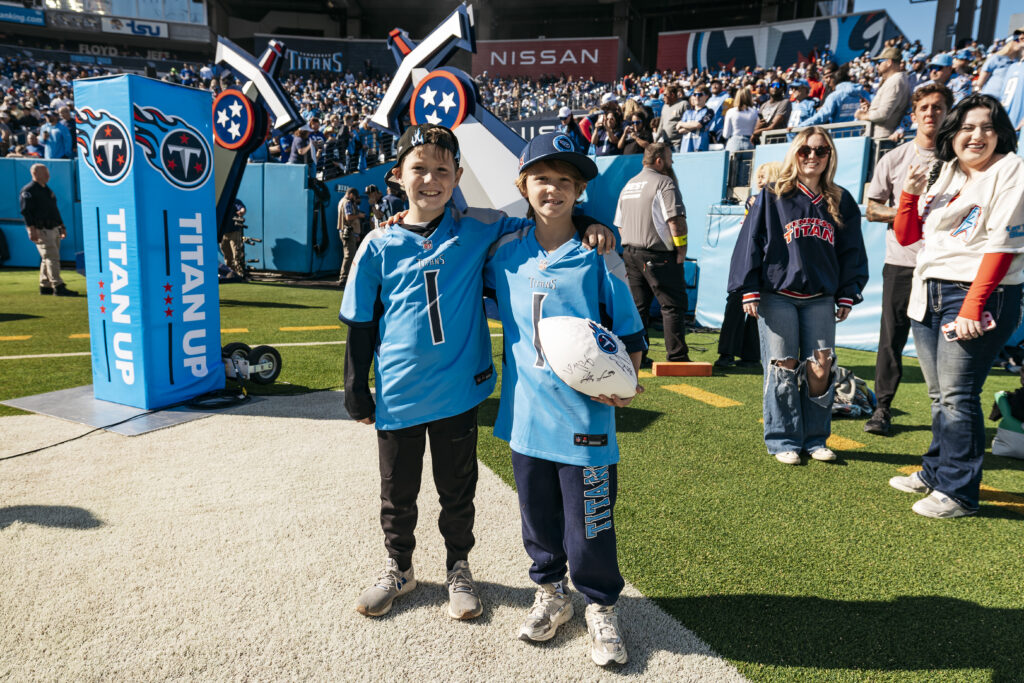 Two smiling children in Tennessee Titans jerseys stand on a football field, one holding a signed football, with fans and stadium seats in the background. It is a sunny day with a lively crowd.