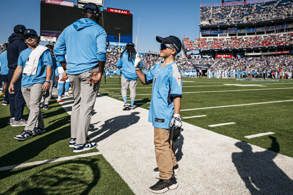 A young boy wearing sunglasses and a light blue football jersey stands on a football field sideline, holding a glove and looking up at an adult. Players and staff fill the background, with a stadium full of fans behind them.