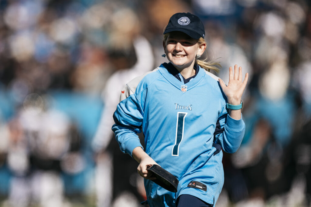 A smiling girl in a blue Tennessee Titans jersey and cap waves while standing on a football field. The background shows a blurred crowd and other players.