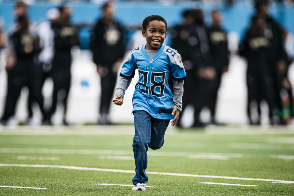 A young boy wearing a blue Tennessee Titans jersey with the number 98 runs joyfully on a football field, with people and blurred figures visible in the background.