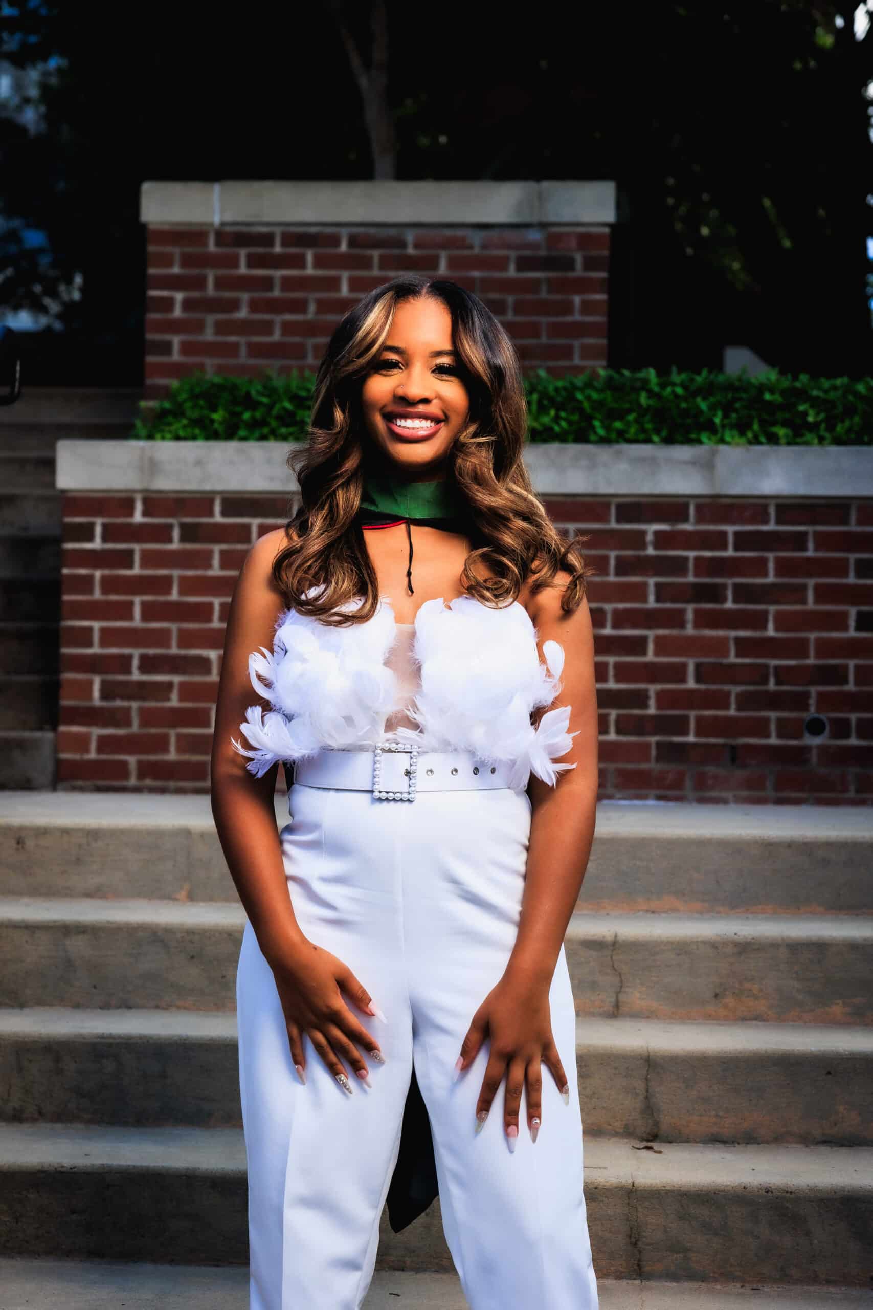 A young woman stands outdoors on stone steps, smiling. She wears white pants and a white top with decorative floral accents, with a green and black graduation stole around her neck. Brick and greenery are in the background.