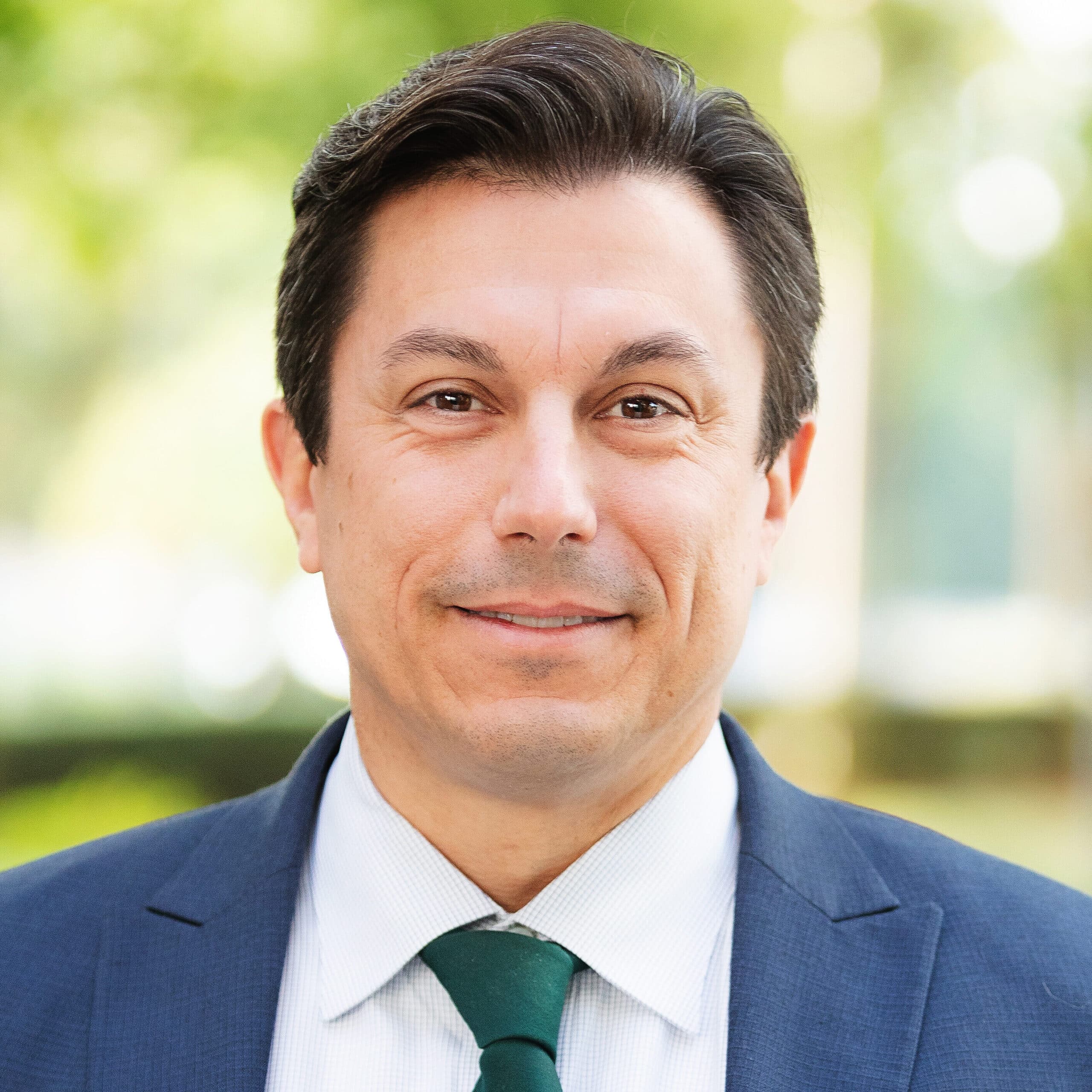A man in a blue suit, white shirt, and green tie smiles while standing outdoors, with a blurred green background of trees and sunlight.