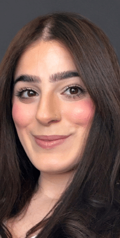 A woman with long dark brown hair and thick eyebrows smiles slightly. She has fair skin, rosy cheeks, and wears subtle makeup. The background is plain and dark.