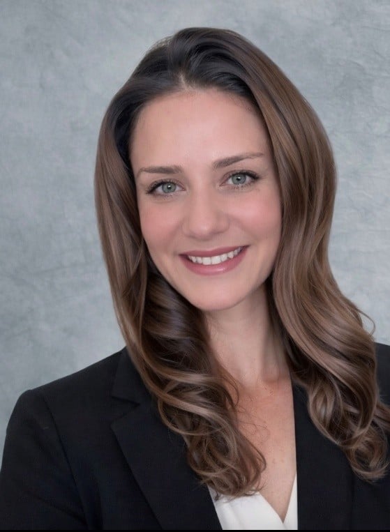 A woman with long brown hair wearing a black blazer and white blouse smiles at the camera. The background is a textured light gray.