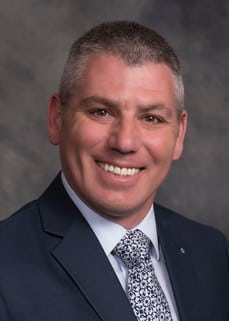 Smiling man with short gray hair wearing a dark suit, white shirt, and patterned tie, posing in front of a gray studio background.