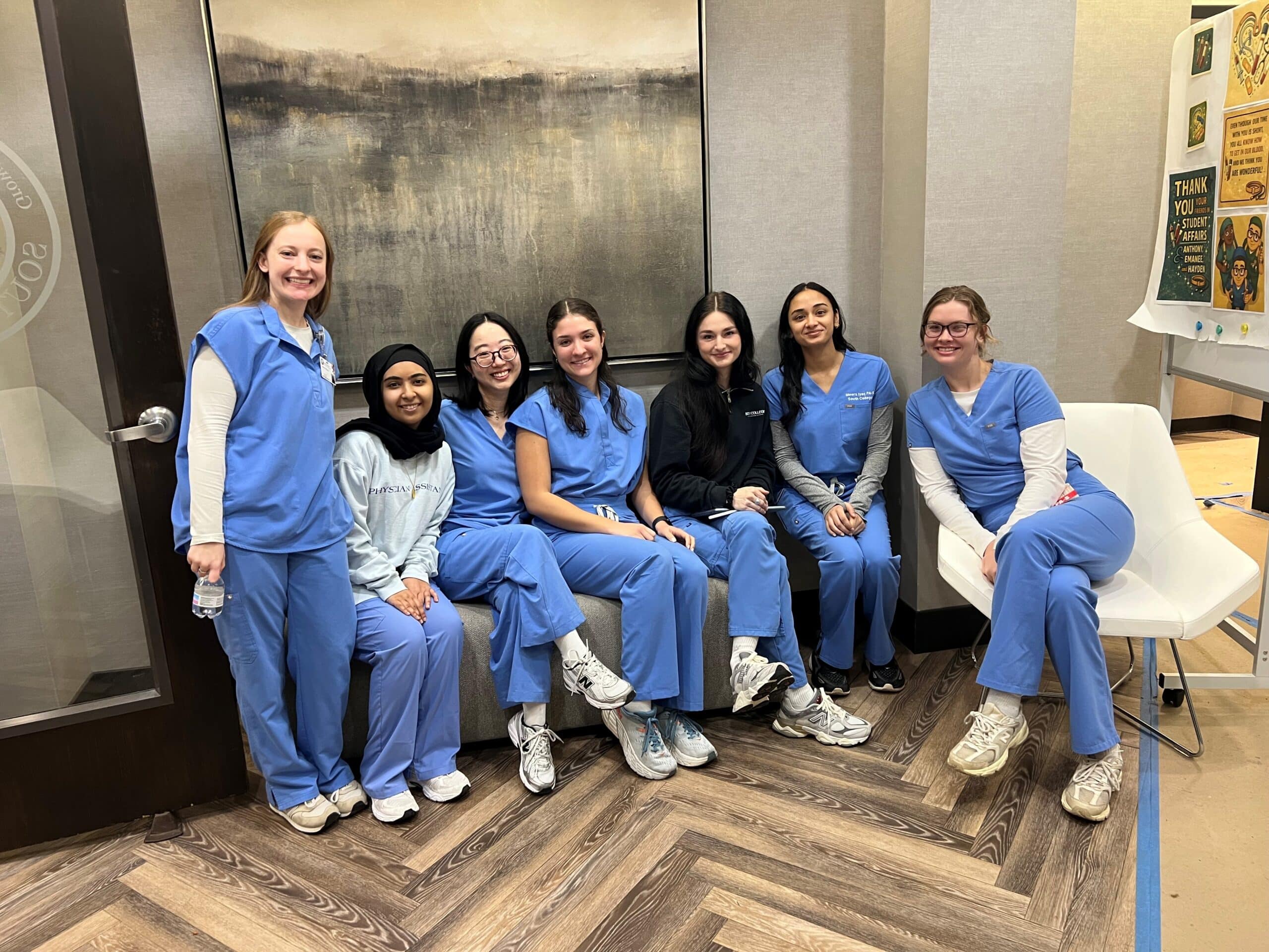 Seven women in blue medical scrubs sit and smile together in a clinic or hospital lounge area. They are seated on a white couch and chair, with a modern painting and informational posters in the background.