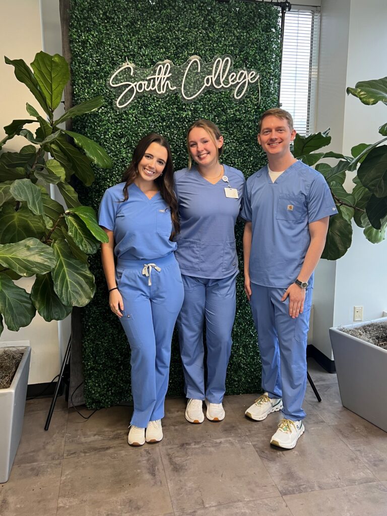 Three people in blue medical scrubs stand smiling in front of a green wall with a "South College" sign. Large potted plants are on either side, and sunlight comes through a window in the background.