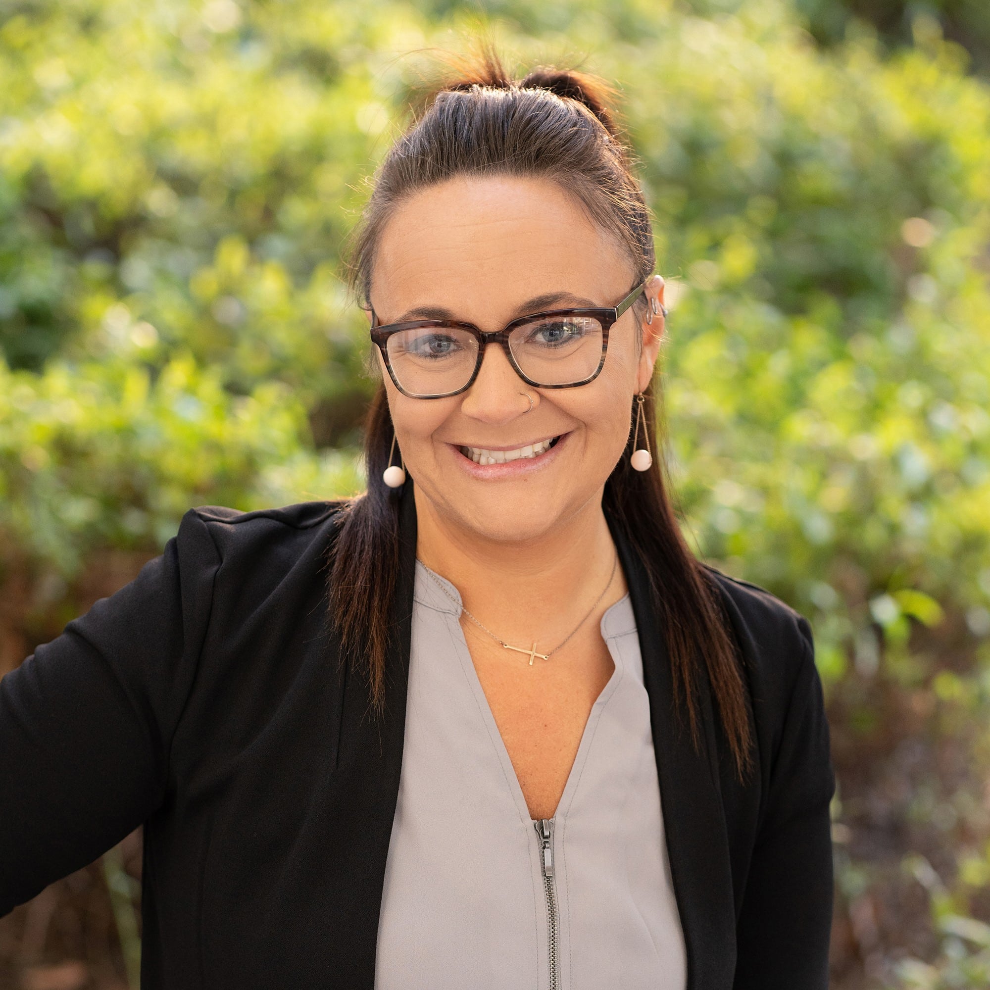 A woman with long brown hair in a ponytail, wearing glasses, a black blazer, and a light gray top, smiles outdoors with greenery in the blurred background.