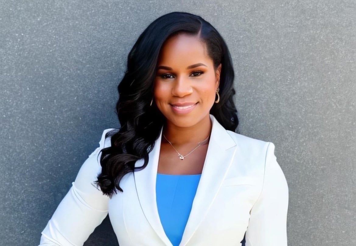 A woman with long, wavy black hair wears a white blazer over a blue top, smiling and standing against a textured gray background. She has hoop earrings and a delicate necklace.
