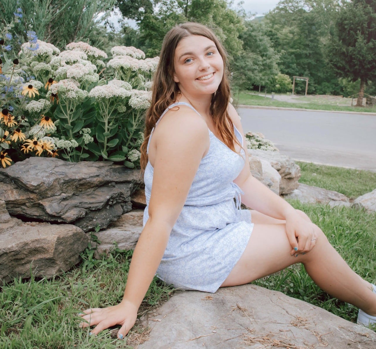 A young woman in a light blue romper sits on a large rock near blooming flowers and greenery, smiling at the camera with a relaxed and cheerful expression. A road and trees are in the background.