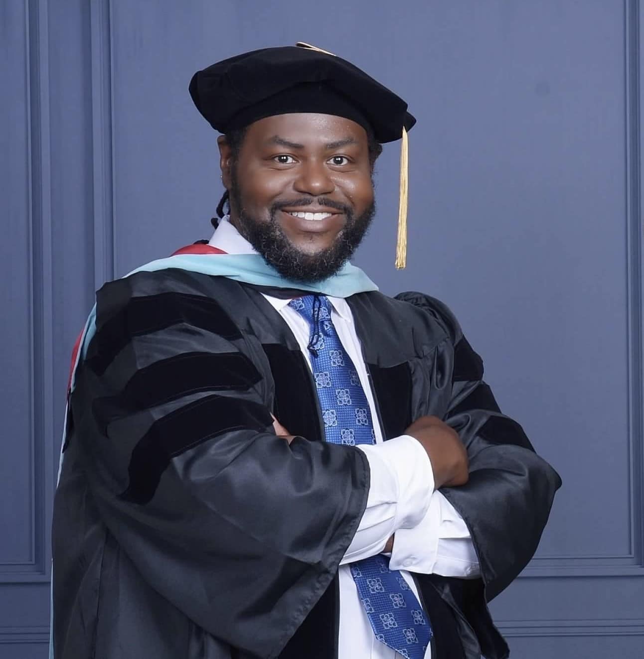 A man wearing academic regalia, including a black graduation cap with a gold tassel and a black gown with velvet panels, stands smiling with arms crossed in front of a blue-gray background.