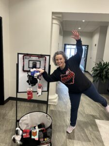 A person in a black "Buckeyes" shirt poses playfully, pretending to dunk socks into a small basketball hoop attached to a stand. A bin of socks and toiletries sits on the floor nearby in a hallway.