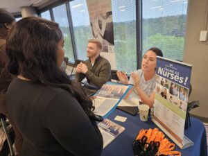A woman speaks to two recruiters at a table with a “Calling All Nurses!” sign, brochures, and orange lanyards, at a career fair near large windows.