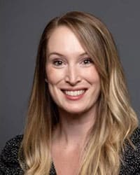 A woman with long, blonde hair and a side part smiles at the camera. She is wearing a black patterned top and is posed in front of a plain gray background.
