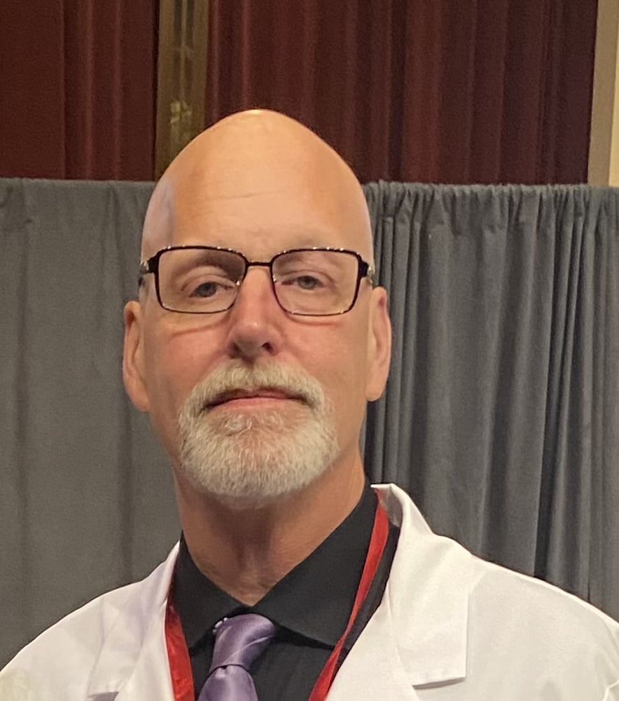 A middle-aged man with glasses and a white goatee is wearing a white coat, black shirt, and purple tie. Gray curtains and maroon walls are visible in the background.