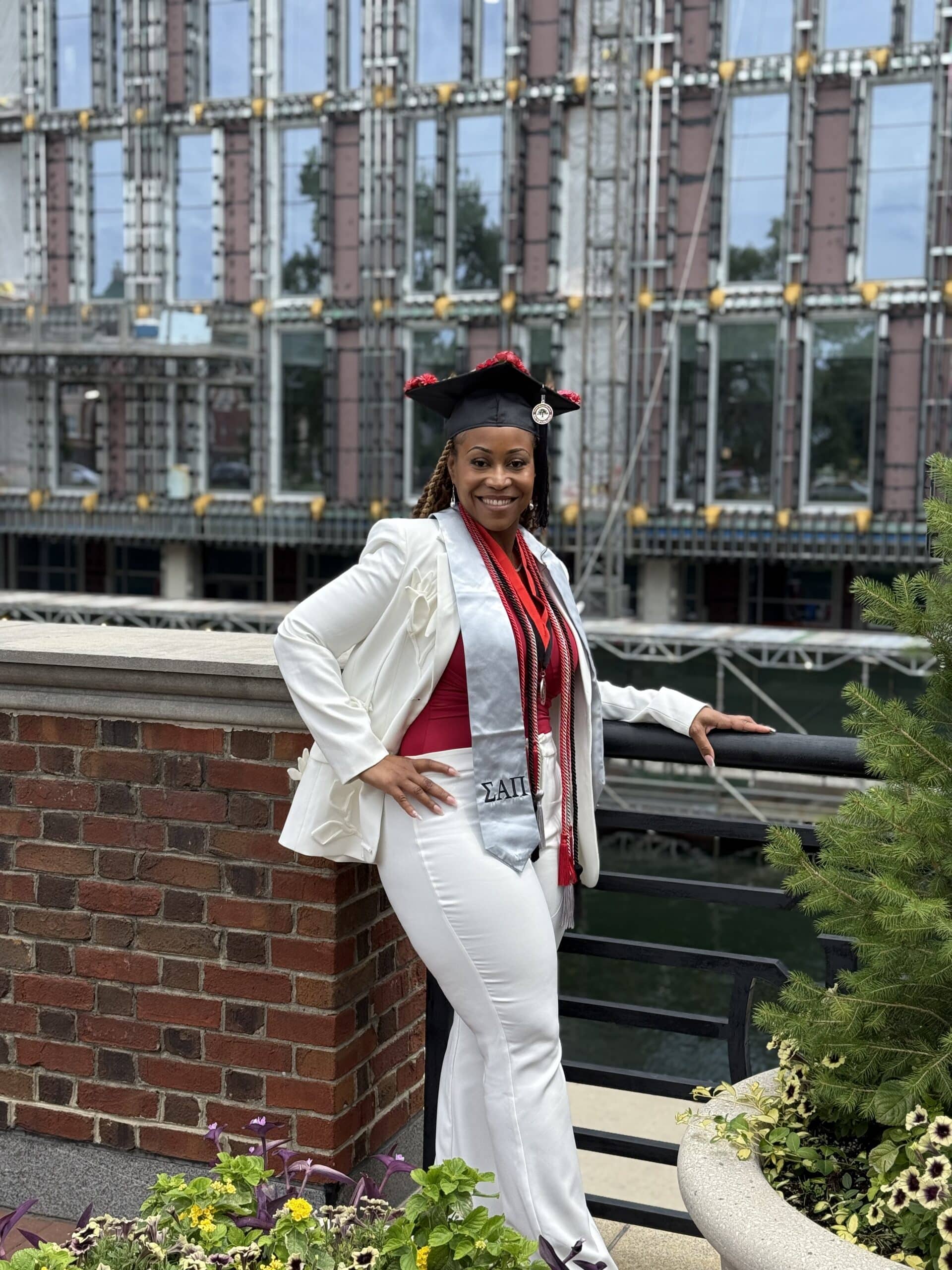 A woman in a white graduation outfit and cap poses, smiling, by a brick and metal railing with a modern building and water in the background. She is wearing a red top, a sash, and several cords around her neck.