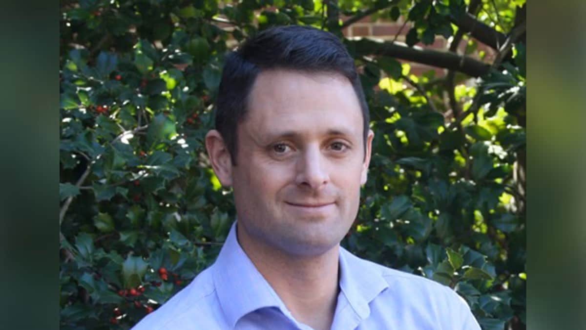 A man with short dark hair, wearing a light blue collared shirt, stands outdoors in front of green leafy bushes with red berries. Sunlight filters through the foliage.