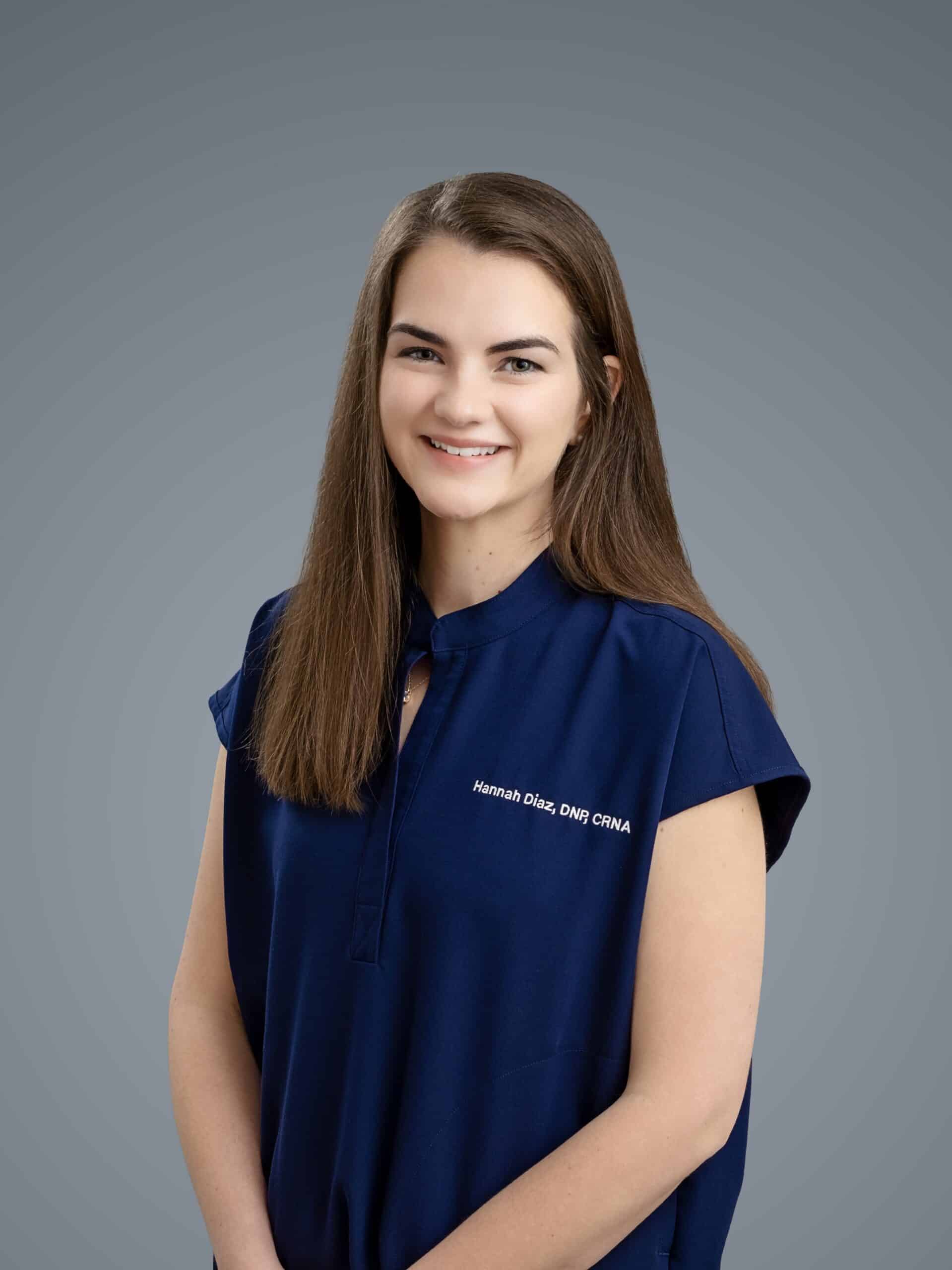 A woman with long brown hair wearing a dark blue shirt that has white text reading "Hannah Diaz, DNP, CRNA" stands against a plain grey background, smiling at the camera.