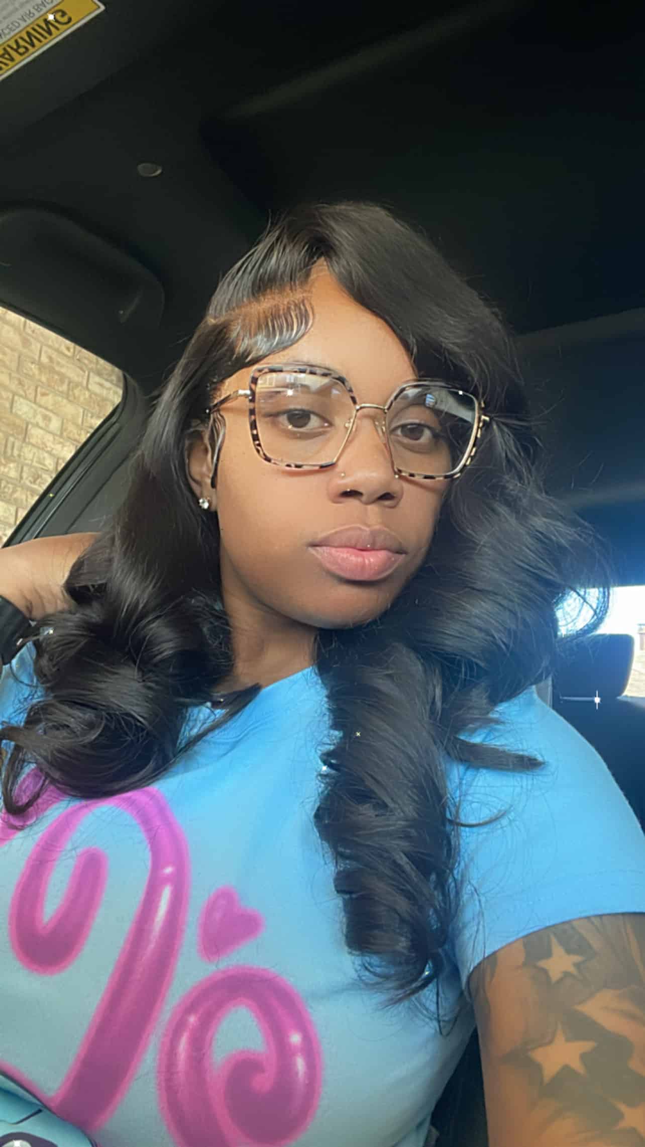 A woman with wavy black hair and large tortoiseshell glasses takes a selfie. She is wearing a blue shirt with pink writing.