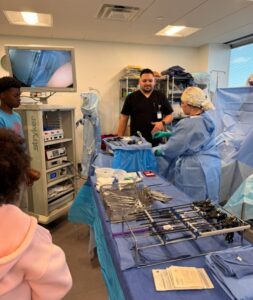 Medical staff in scrubs and surgical masks stand beside a table with surgical instruments, conducting a demonstration. A monitor displays an internal view. Two observers watch in the foreground. The room is well-lit with large windows.