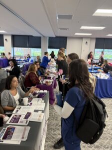 A group of people interact at a busy indoor career or resource fair, with tables covered in brochures, flyers, and promotional items. Attendees, including some in scrubs, speak to representatives behind the tables.