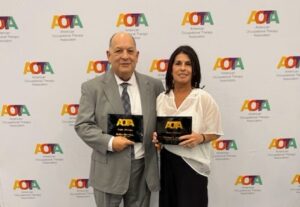 Two people stand side by side holding plaques in front of an "AOTA" (American Occupational Therapy Association) step-and-repeat banner, both dressed formally and smiling at the camera.