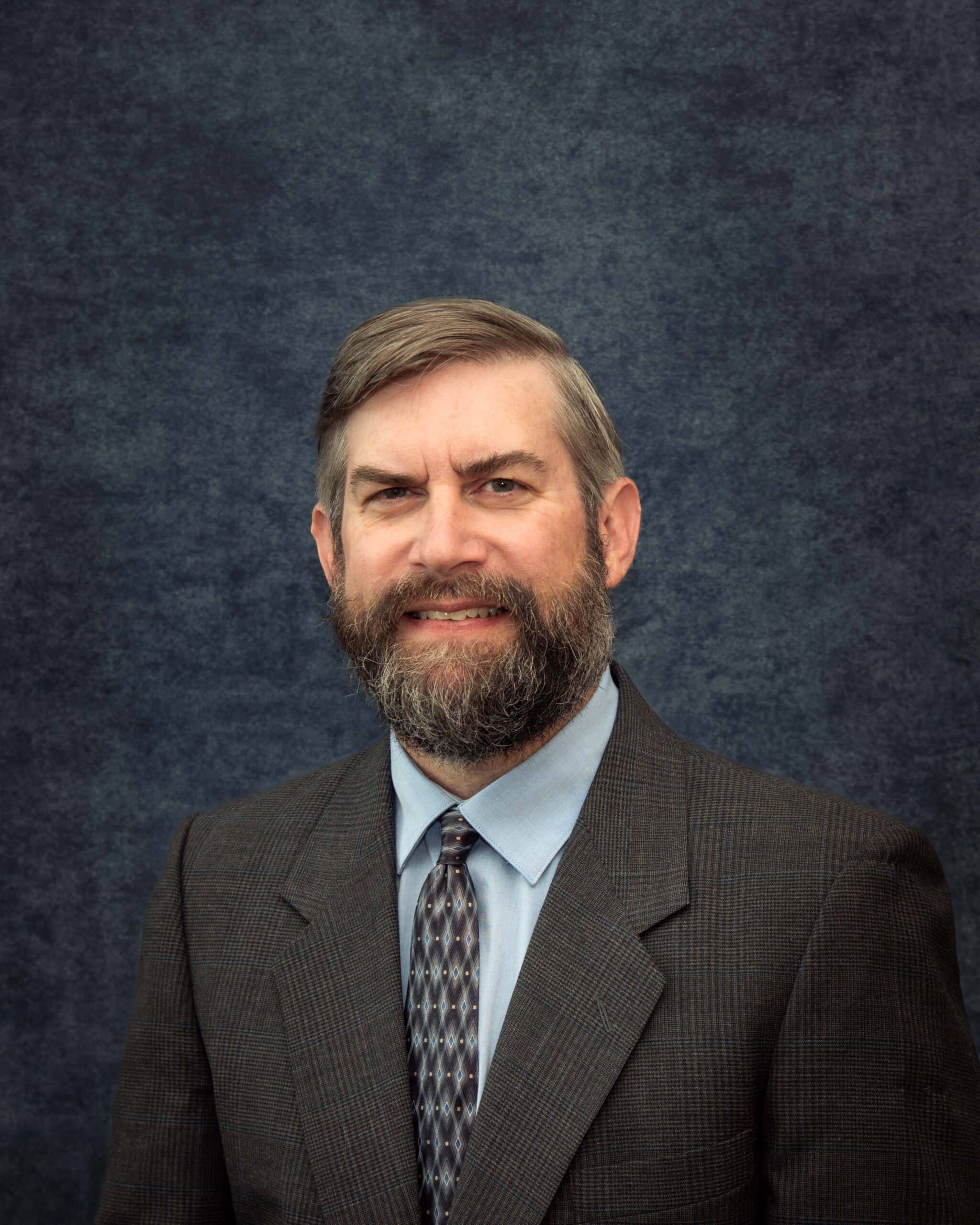 A man with short brown hair and a full beard is wearing a dark suit jacket, light blue shirt, and patterned tie, posing in front of a dark, textured backdrop.