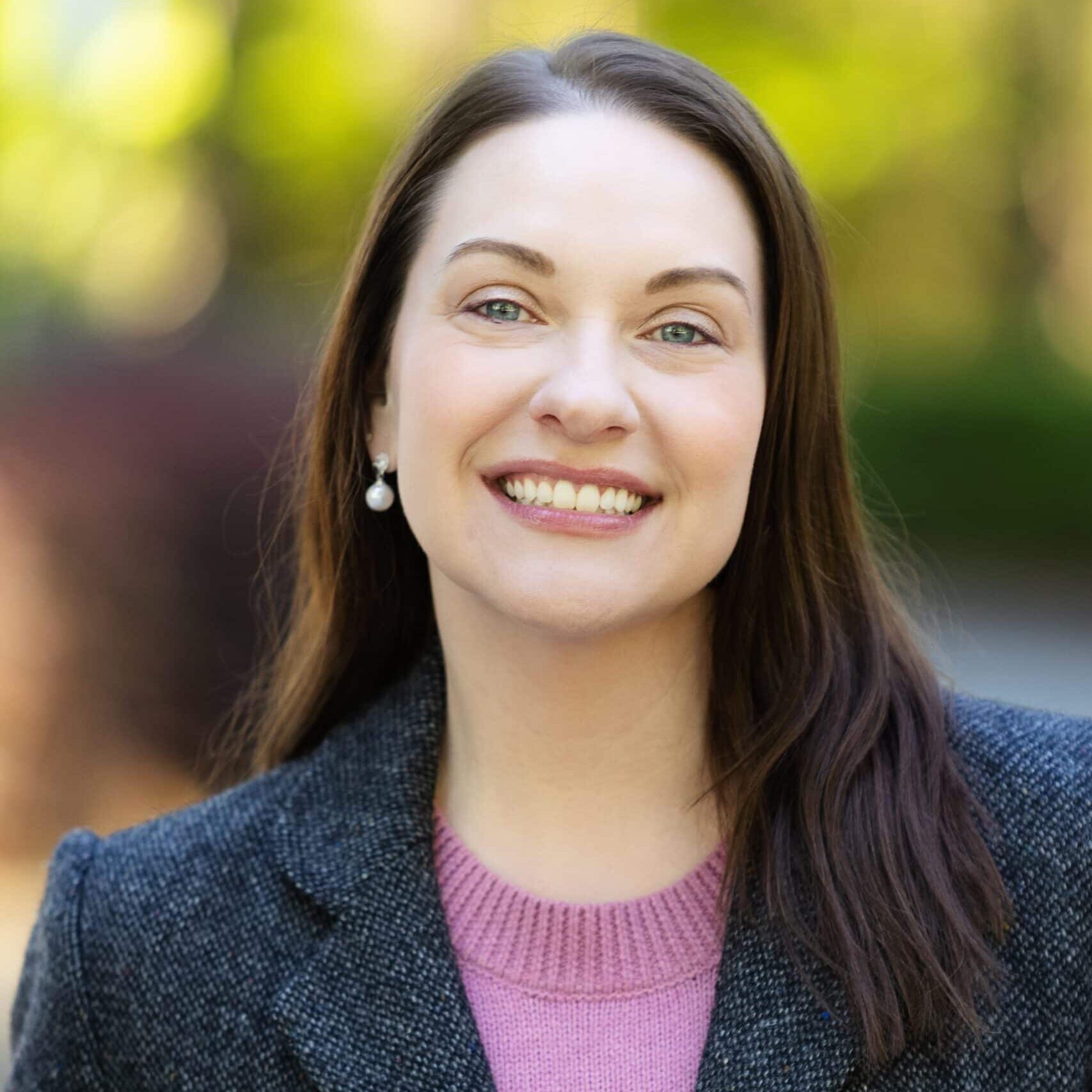 A woman with straight brown hair, wearing a gray blazer over a pink sweater, smiles at the camera outdoors with a blurred green and brown background.