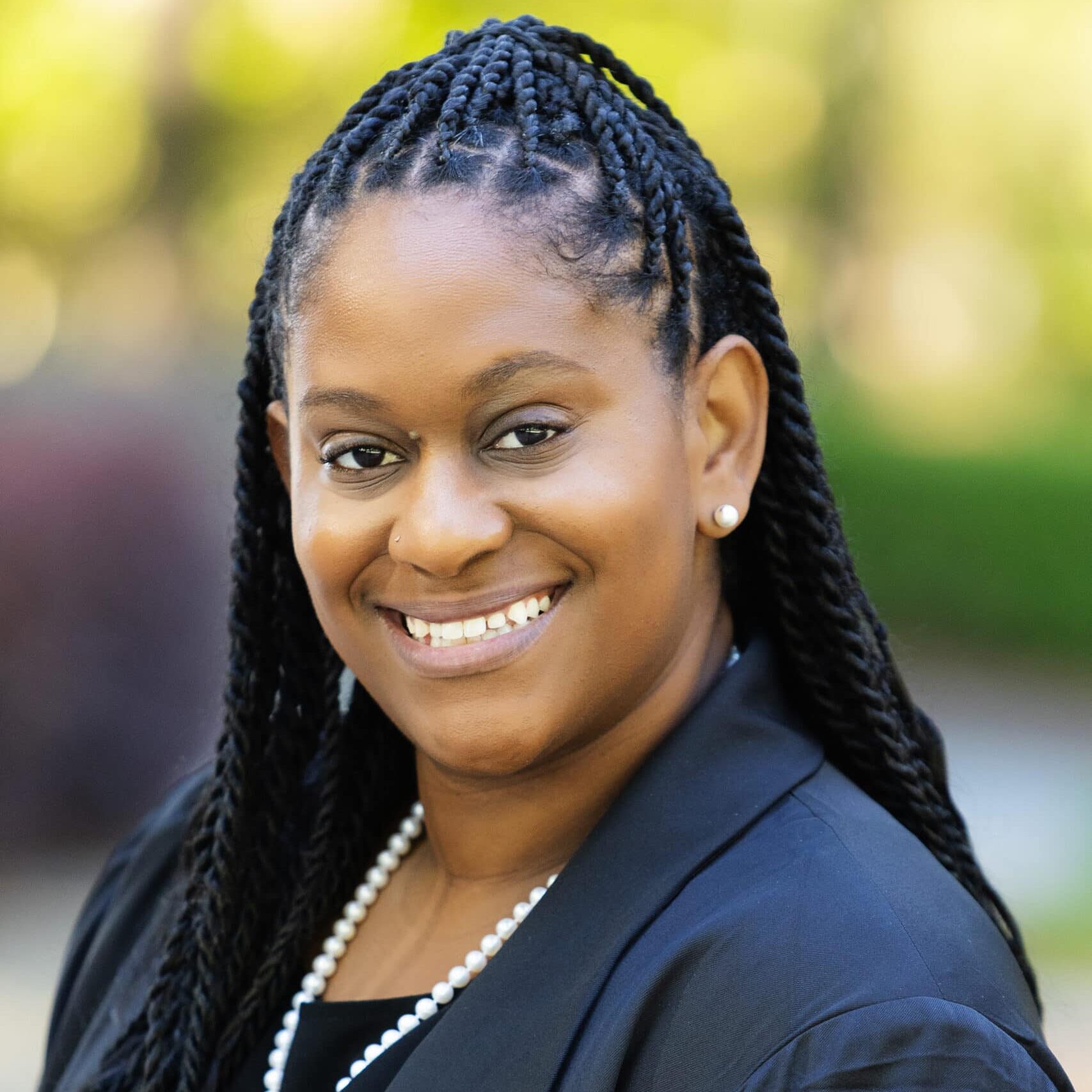 A woman with long braided hair, wearing a black blazer, a black top, and a pearl necklace, smiles at the camera. The background is blurred greenery and sunlight.