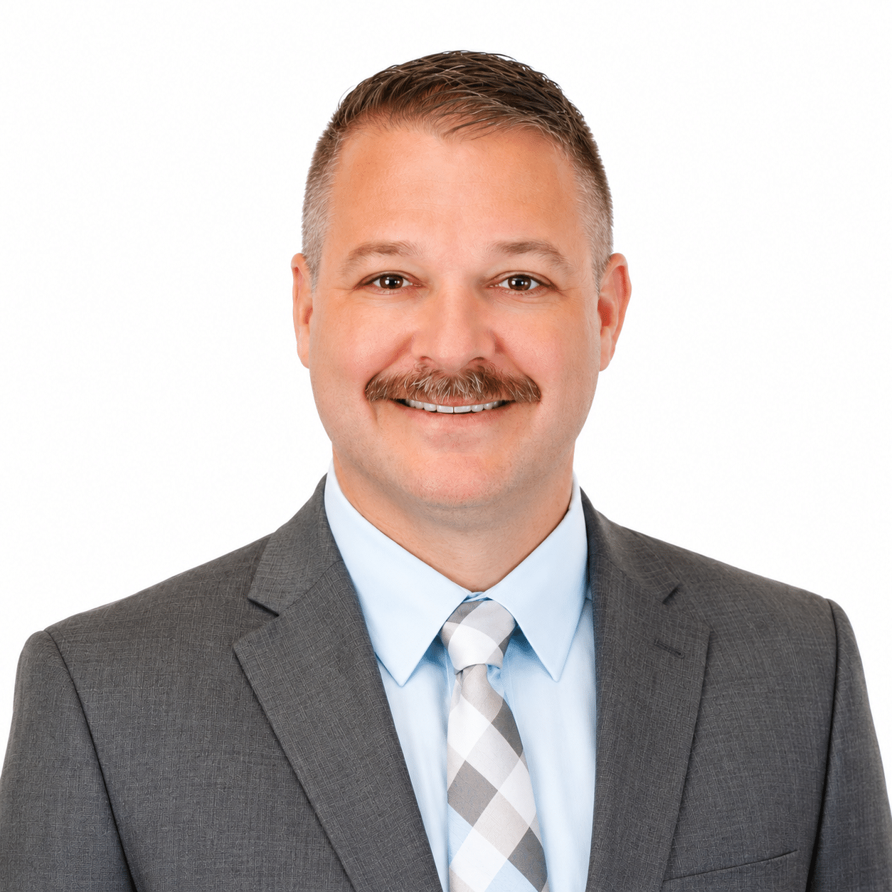 A man in a gray suit, light blue shirt, and plaid tie smiles at the camera. He has short brown hair, a mustache, and stands against a plain white background.