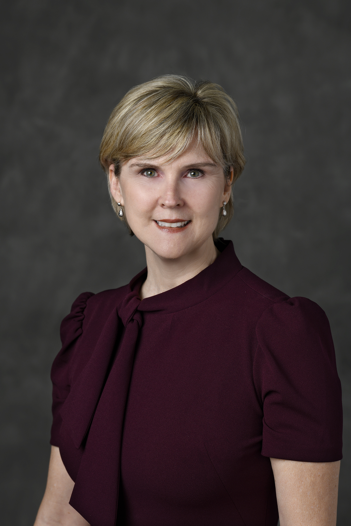 A woman with short blonde hair wearing a burgundy top with a bow detail, smiling and posing in front of a gray studio background.