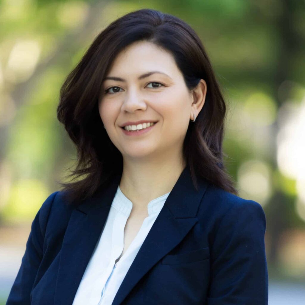 A woman with shoulder-length dark hair, wearing a navy blazer and white blouse, stands outdoors smiling with a blurred background of greenery.