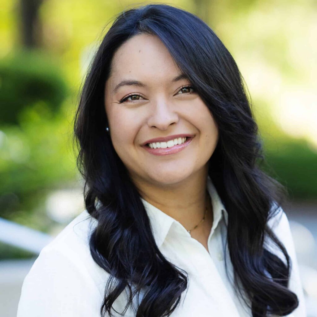 A woman with long, wavy dark hair smiles at the camera. She is wearing a white blouse and is outdoors, with greenery and sunlight blurred in the background.