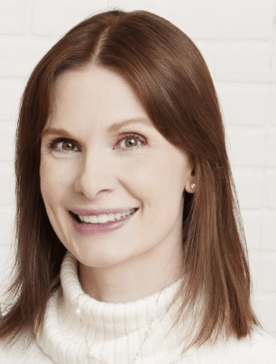 A woman with straight, shoulder-length brown hair smiles at the camera. She is wearing a white turtleneck sweater and a delicate necklace, standing in front of a white brick wall.