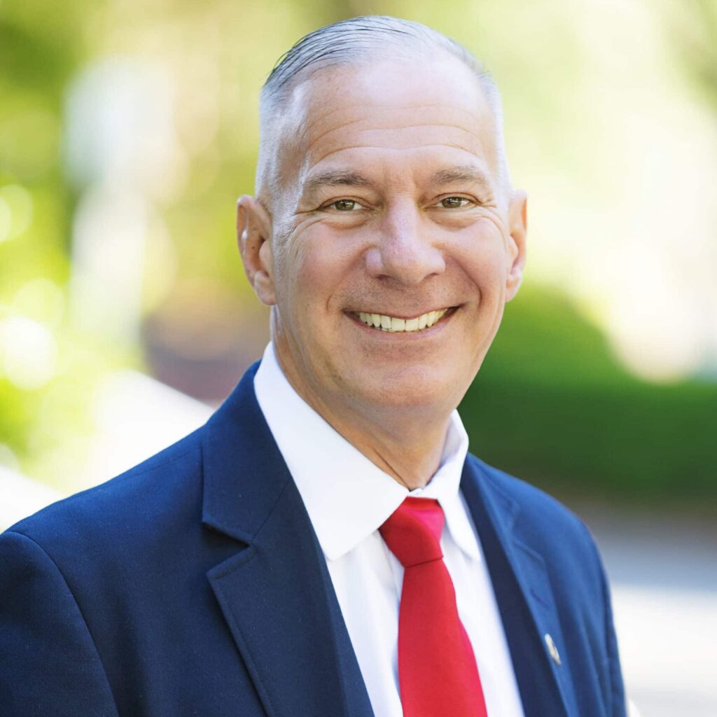 A smiling man with short gray hair wears a dark suit, white shirt, and bright red tie. He stands outdoors with a blurred, green background.