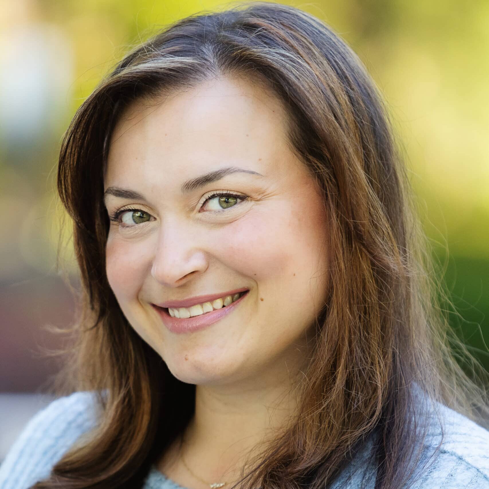 A woman with long brown hair and light eyes smiles at the camera. She is wearing a light gray ribbed sweater and is outdoors with a blurred green background.