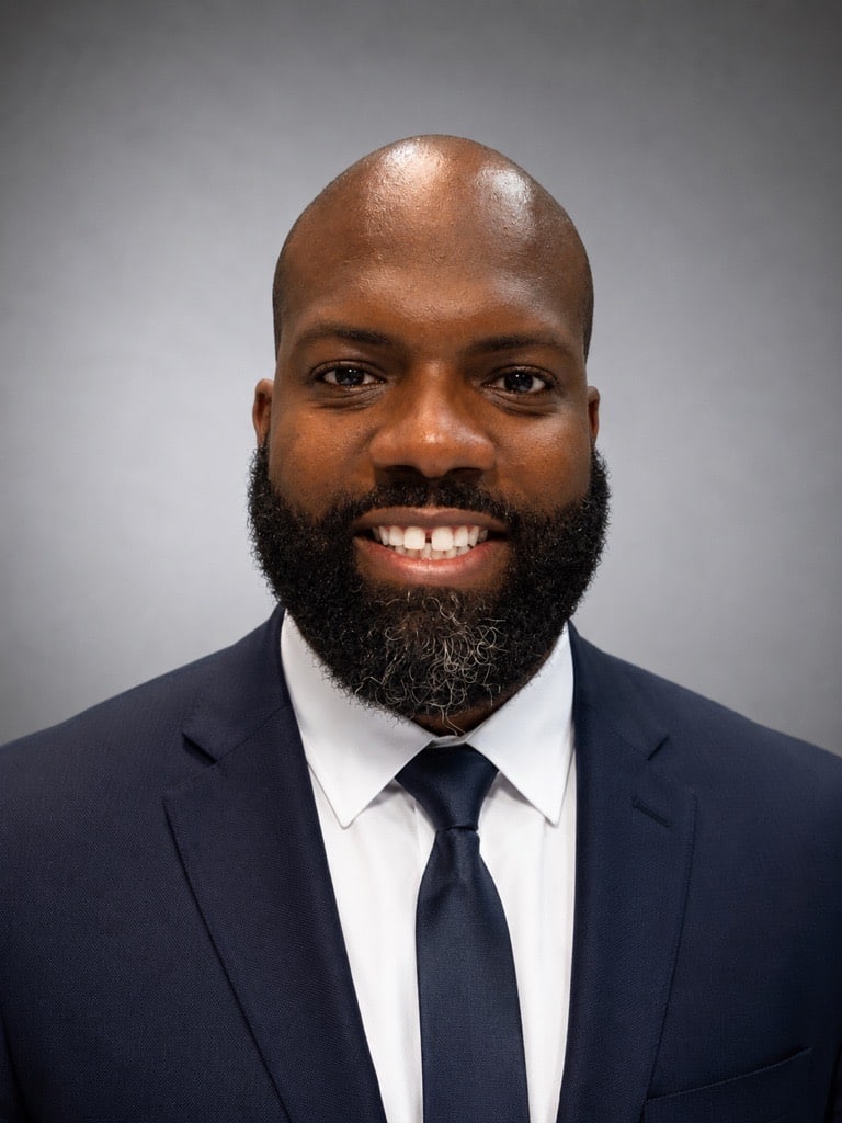 A man with a full beard and mustache, wearing a dark blue suit, white dress shirt, and dark tie, smiles at the camera against a plain gray background.