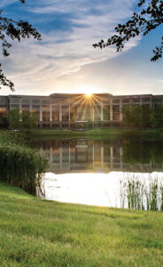 A modern building labeled "South College" stands behind a reflective pond at sunset, with sun rays shining above the roof, trees framing the scene, and grass in the foreground.