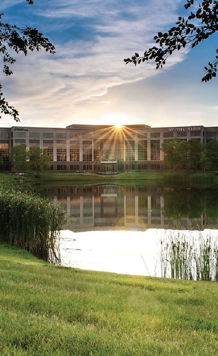 A modern building labeled "South College" stands behind a reflective pond at sunset, with sun rays shining above the roof, trees framing the scene, and grass in the foreground.