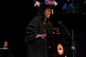 A woman in academic regalia smiles while speaking at a podium during a graduation ceremony. A microphone and a seal with a tree logo are visible on the podium. Other people in academic attire are seated in the background.
