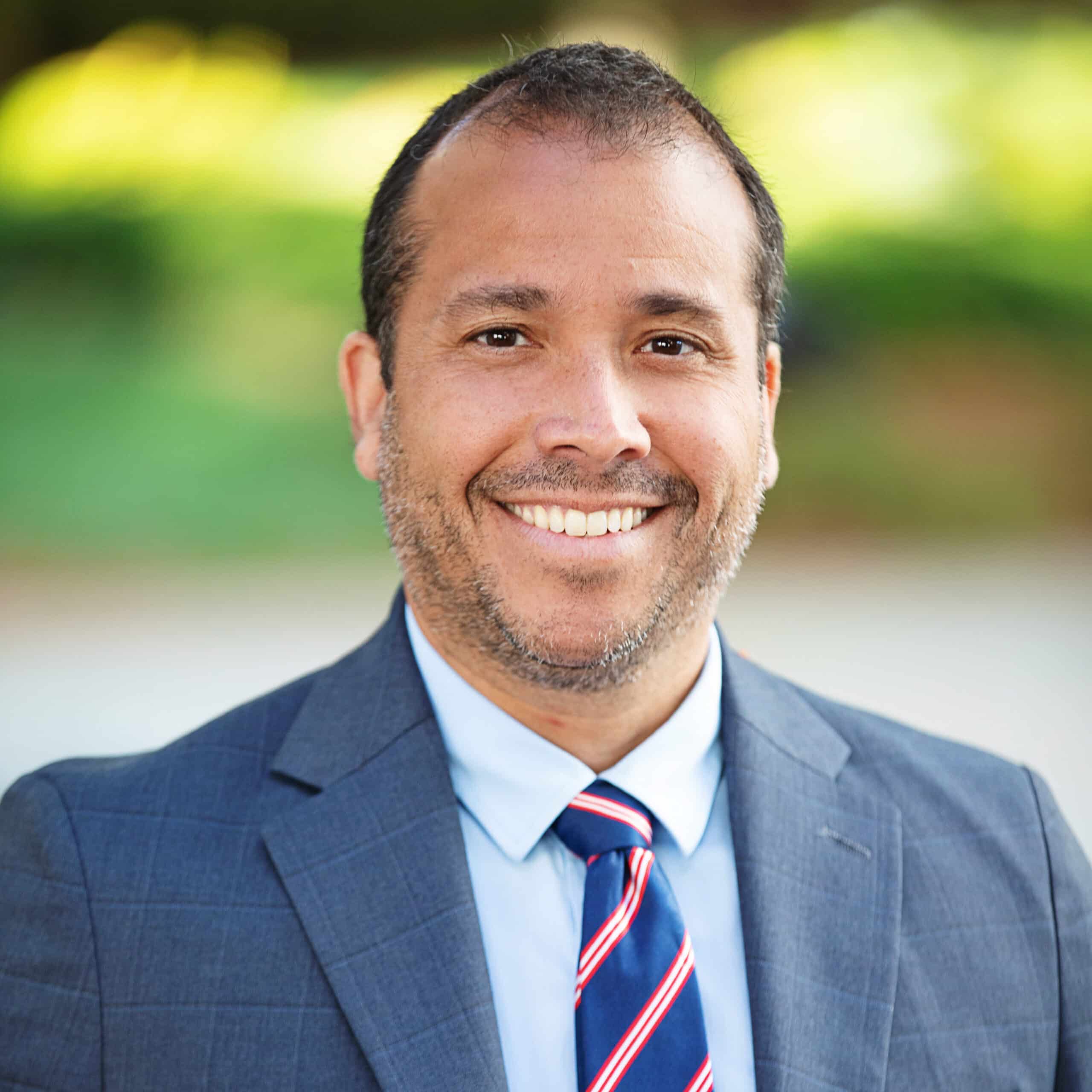 A man with short dark hair and a beard smiles warmly at the camera. He is wearing a blue suit, a light blue shirt, and a blue tie with red and white stripes. The background is blurred with green foliage, suggesting an outdoor setting.