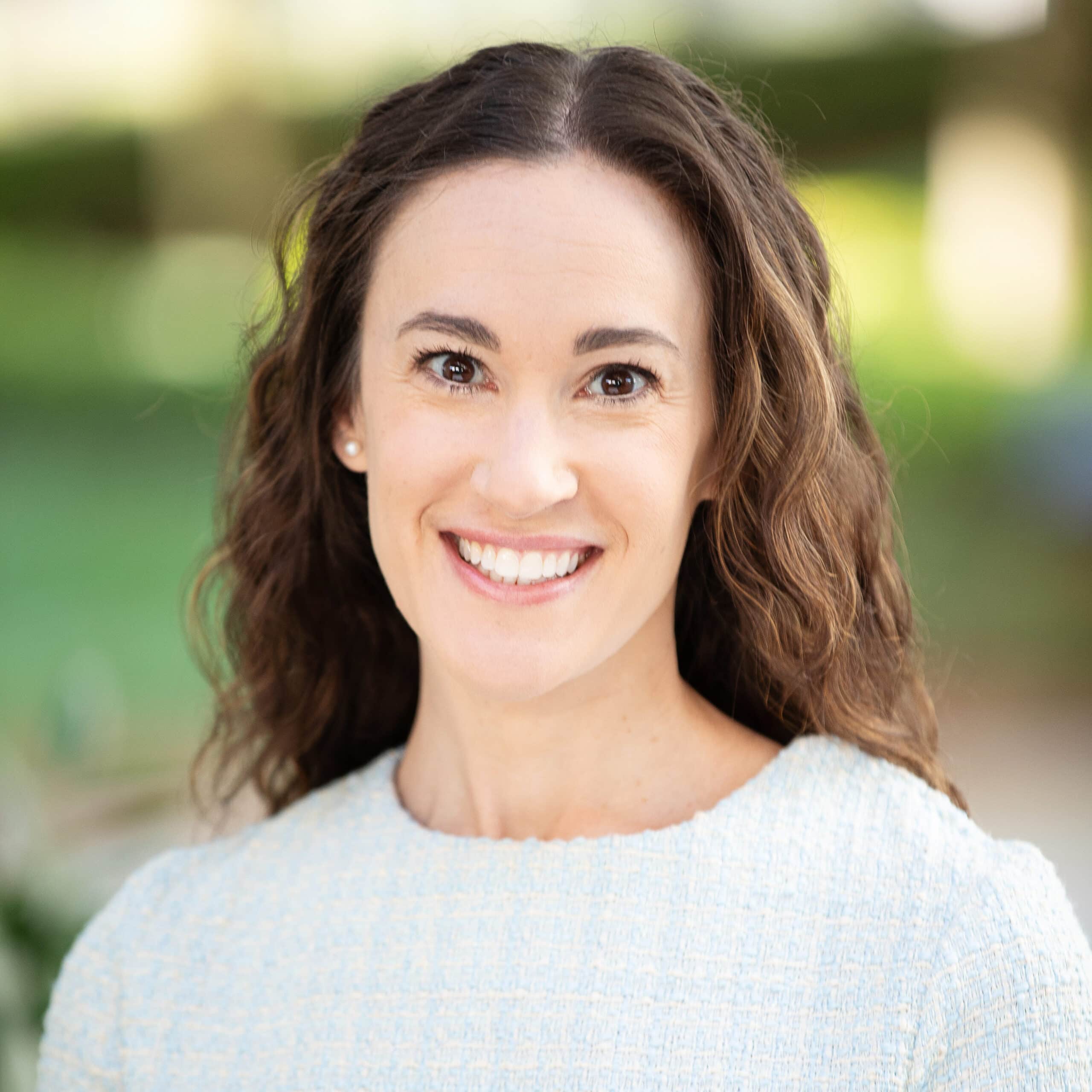 A woman with long, curly brown hair is smiling at the camera. She is wearing a light blue short-sleeved top and standing outdoors with a blurred green background.