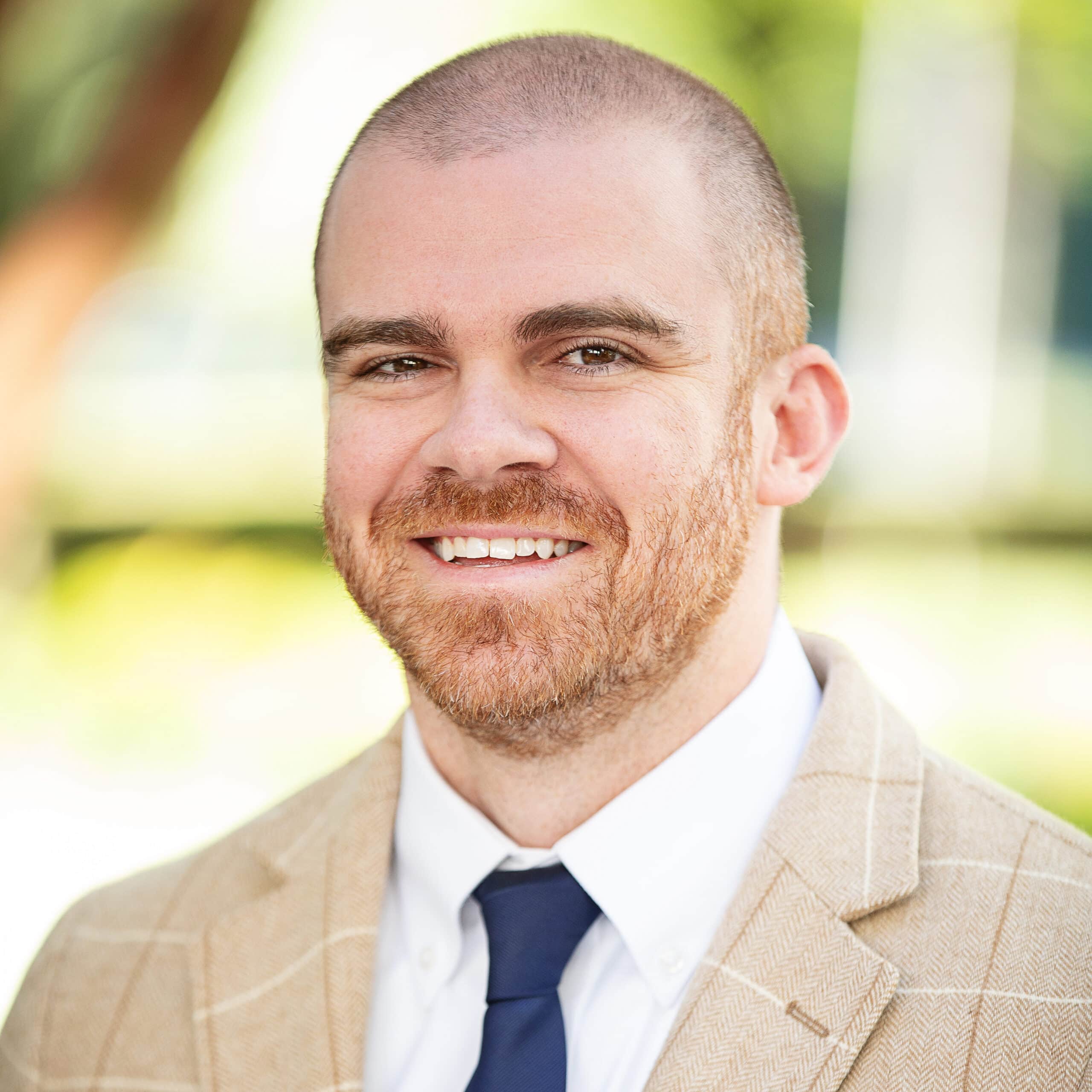 A man with a beard and a shaved head is wearing a beige plaid suit jacket, a white dress shirt, and a dark blue tie. He is smiling and the background is softly blurred with greenery.