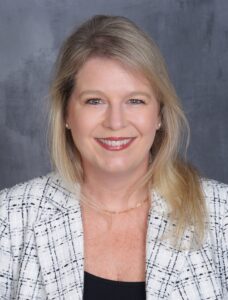 A woman with long blonde hair wearing a white patterned blazer and a black top smiles at the camera against a gray background.