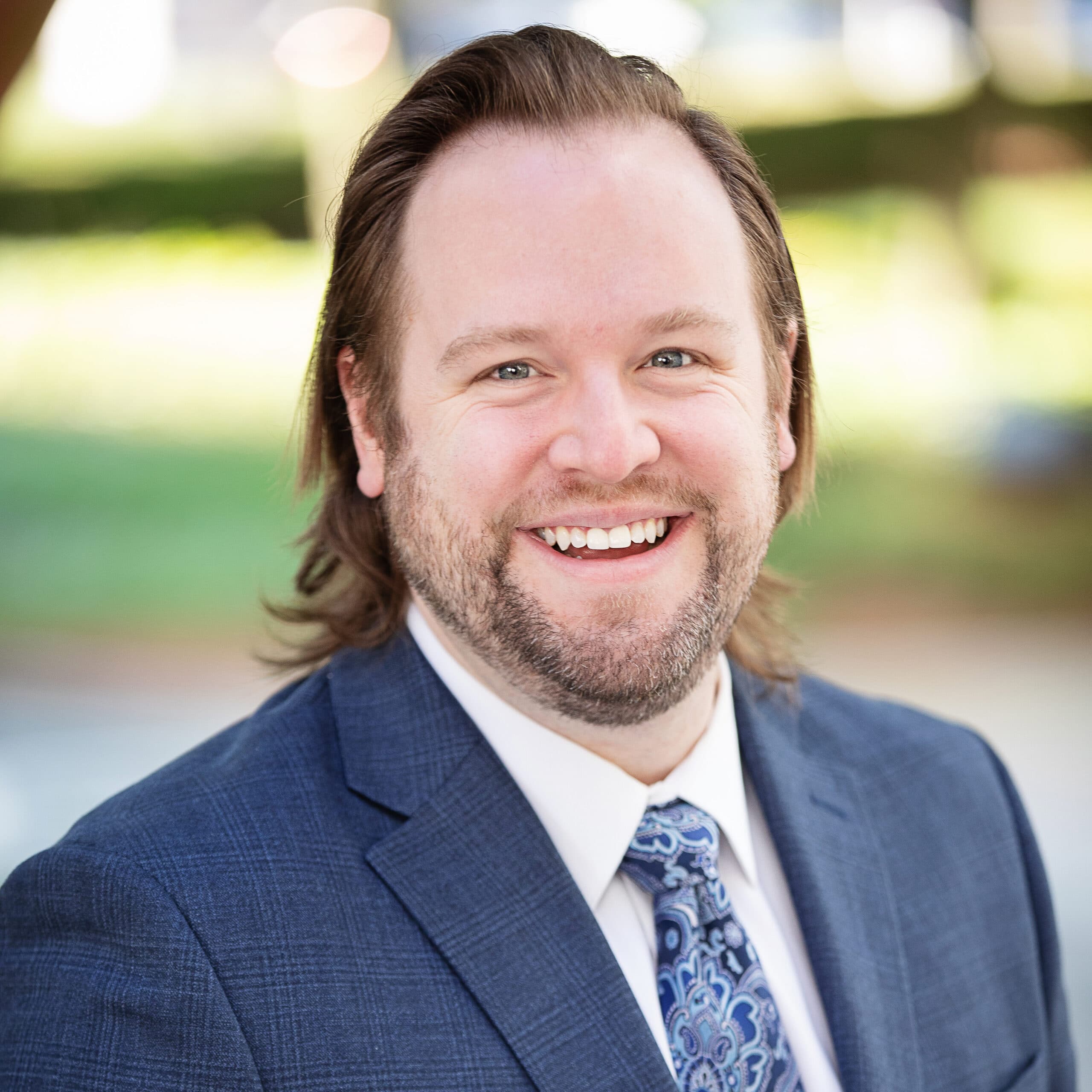 A bearded man with shoulder-length hair smiles warmly at the camera. He is wearing a blue suit jacket, white dress shirt, and a blue paisley tie. The background is blurred with greenery and outdoor elements visible.
