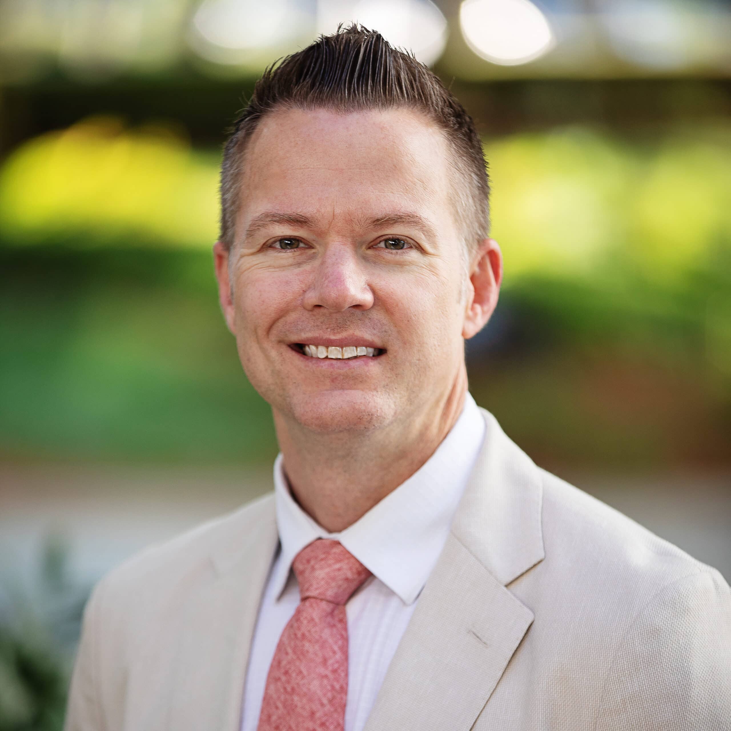 A man is smiling and wearing a light beige suit with a white shirt and a peach-colored tie. He has short, neatly styled hair. The background is outdoors with greenery and blurred elements.