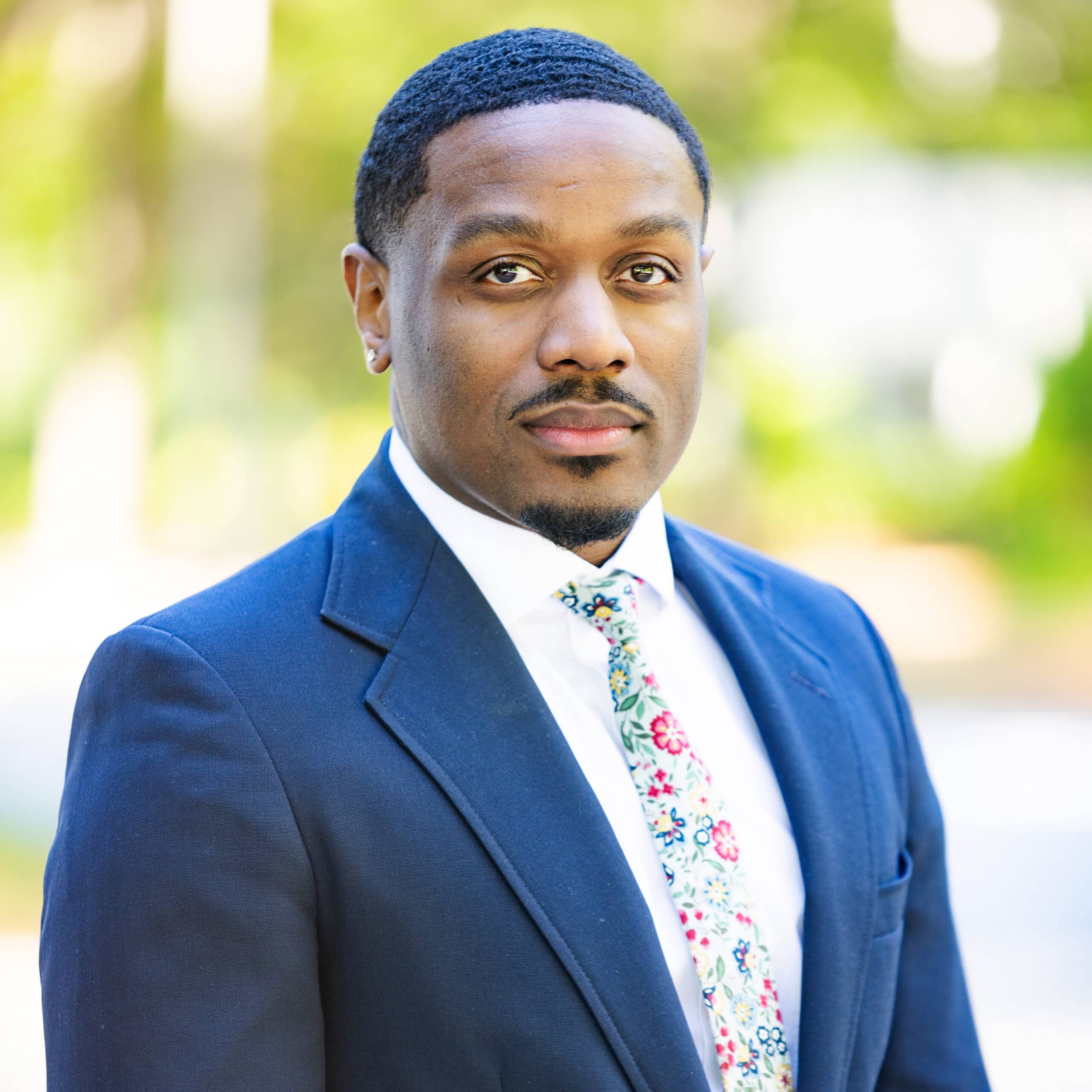 A man wearing a navy blue suit, white shirt, and floral-patterned tie stands outdoors with a blurred green and white background, looking confidently at the camera.